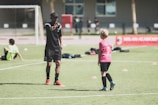 A soccer coach instructs a young player on a field set up with orange cones. The coach, dressed in black with red shoes, gestures towards the player in a pink shirt and black shorts. Other children in sports attire are visible in the background, with a soccer net and a red banner for the Milan Academy nearby.