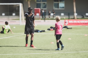 A soccer coach instructs a young player on a field set up with orange cones. The coach, dressed in black with red shoes, gestures towards the player in a pink shirt and black shorts. Other children in sports attire are visible in the background, with a soccer net and a red banner for the Milan Academy nearby.