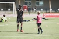A soccer coach instructs a young player on a field set up with orange cones. The coach, dressed in black with red shoes, gestures towards the player in a pink shirt and black shorts. Other children in sports attire are visible in the background, with a soccer net and a red banner for the Milan Academy nearby.