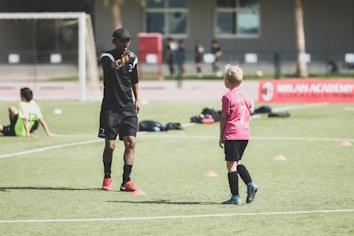 A soccer coach instructs a young player on a field set up with orange cones. The coach, dressed in black with red shoes, gestures towards the player in a pink shirt and black shorts. Other children in sports attire are visible in the background, with a soccer net and a red banner for the Milan Academy nearby.