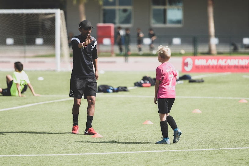 A soccer coach instructs a young player on a field set up with orange cones. The coach, dressed in black with red shoes, gestures towards the player in a pink shirt and black shorts. Other children in sports attire are visible in the background, with a soccer net and a red banner for the Milan Academy nearby.
