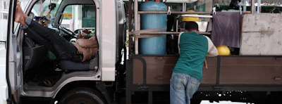 A man rests with his legs up inside the cabin of a parked small truck while another man leans on the back, which carries gas cylinders and other equipment. Clothing and towels hang from the truck, adding a casual feel.