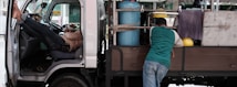 A man rests with his legs up inside the cabin of a parked small truck while another man leans on the back, which carries gas cylinders and other equipment. Clothing and towels hang from the truck, adding a casual feel.