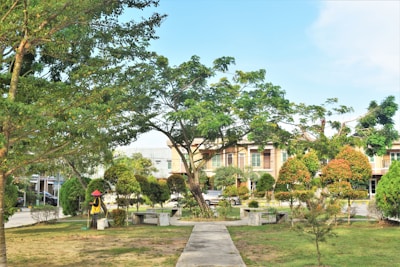 A well-maintained garden area with a paved walkway leading through a grassy field. Various trees and shrubs are arranged around the space, providing a lush, green environment. A figure wearing a red hat and yellow shirt appears to be gardening or maintaining the area. In the background, there are multi-story residential buildings with balconies and windows.
