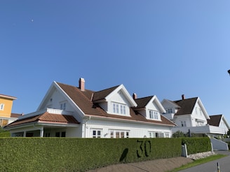 A row of similar white wooden houses with slanted brown tiled roofs are aligned alongside a neatly trimmed green hedge with the number 30 visible. The sky is clear and blue, creating a bright and peaceful suburban setting.