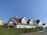 A row of similar white wooden houses with slanted brown tiled roofs are aligned alongside a neatly trimmed green hedge with the number 30 visible. The sky is clear and blue, creating a bright and peaceful suburban setting.