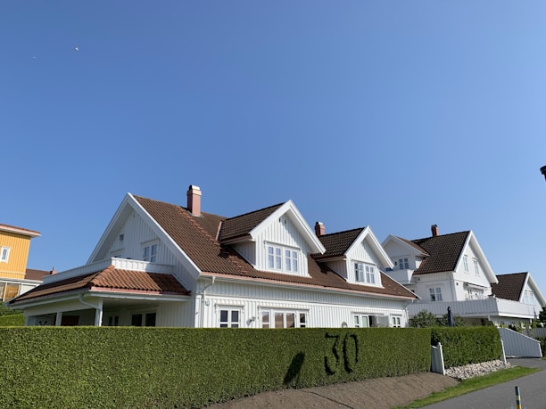 A row of similar white wooden houses with slanted brown tiled roofs are aligned alongside a neatly trimmed green hedge with the number 30 visible. The sky is clear and blue, creating a bright and peaceful suburban setting.