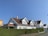 A row of similar white wooden houses with slanted brown tiled roofs are aligned alongside a neatly trimmed green hedge with the number 30 visible. The sky is clear and blue, creating a bright and peaceful suburban setting.