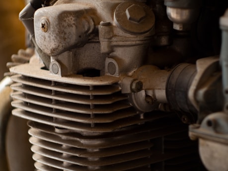 A close-up view of an engine component featuring a weathered, metallic surface with bolts and a series of horizontal cooling fins. The golden-brown hue suggests aging or exposure to environmental elements.