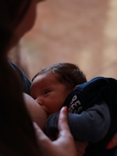 A close-up of a baby breastfeeding, with a soft, intimate focus. The baby's gaze is upward, and the person's hand gently holds the baby, conveying a sense of care and tenderness.