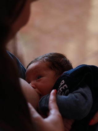 A close-up of a baby breastfeeding, with a soft, intimate focus. The baby's gaze is upward, and the person's hand gently holds the baby, conveying a sense of care and tenderness.