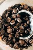 A close-up view of a pile of roasted coffee beans arranged in and around a metal grinder or container. The beans have a rich, dark brown hue and an oily sheen, with visible creases and textures.