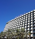 A modern hotel building with a distinctive grid-like facade featuring numerous balconies, and a clear blue sky overhead. Palm trees are visible in the foreground, adding a tropical feel to the scene.