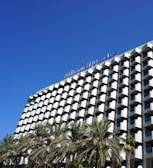 A modern hotel building with a distinctive grid-like facade featuring numerous balconies, and a clear blue sky overhead. Palm trees are visible in the foreground, adding a tropical feel to the scene.