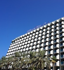 A modern hotel building with a distinctive grid-like facade featuring numerous balconies, and a clear blue sky overhead. Palm trees are visible in the foreground, adding a tropical feel to the scene.