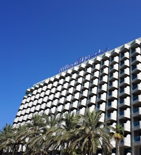 A modern hotel building with a distinctive grid-like facade featuring numerous balconies, and a clear blue sky overhead. Palm trees are visible in the foreground, adding a tropical feel to the scene.