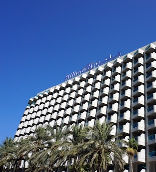 A modern hotel building with a distinctive grid-like facade featuring numerous balconies, and a clear blue sky overhead. Palm trees are visible in the foreground, adding a tropical feel to the scene.