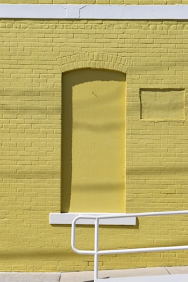 A yellow brick wall featuring a bricked-up doorway under an arch and a square patch. The wall is accented with a white border at the top and a white handrail in the foreground, creating geometric contrasts.