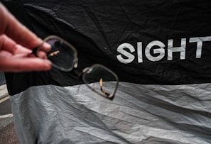 A hand holding a pair of eyeglasses, with a background featuring a black and white fabric with the word 'SIGHT' printed on it.