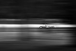 A sleek race car is speeding along a track, captured in black and white. The motion blur effect emphasizes the high speed of the vehicle, with the background and foreground lines appearing stretched and dynamic.