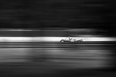 A sleek race car is speeding along a track, captured in black and white. The motion blur effect emphasizes the high speed of the vehicle, with the background and foreground lines appearing stretched and dynamic.