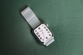 A wristwatch with a white face and black numerals is displayed on a textured green surface. The watch has a silver metal mesh strap and features minimalist design elements, including simple hour and minute hands.
