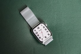 A wristwatch with a white face and black numerals is displayed on a textured green surface. The watch has a silver metal mesh strap and features minimalist design elements, including simple hour and minute hands.