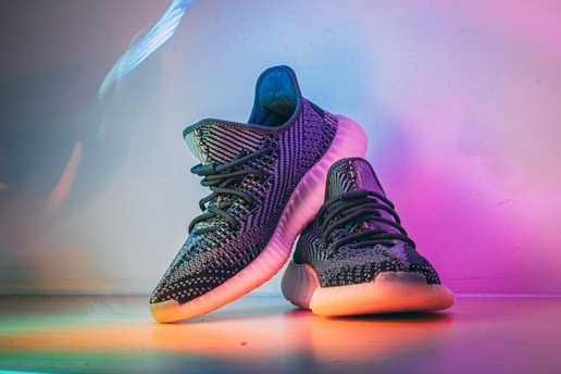 A pair of stylish, dark-colored sneakers with textured patterns, placed on a surface that reflects ambient rainbow-colored lighting. The shoes have a distinct sole design, prominent laces, and exhibit a modern, fashionable look.