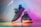 A pair of stylish, dark-colored sneakers with textured patterns, placed on a surface that reflects ambient rainbow-colored lighting. The shoes have a distinct sole design, prominent laces, and exhibit a modern, fashionable look.