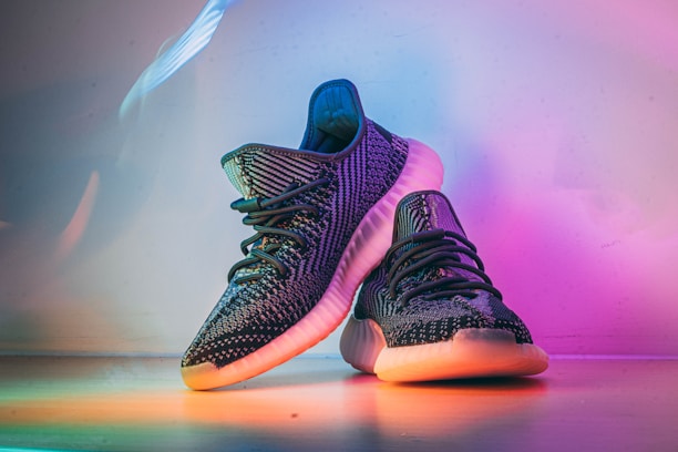 A pair of stylish, dark-colored sneakers with textured patterns, placed on a surface that reflects ambient rainbow-colored lighting. The shoes have a distinct sole design, prominent laces, and exhibit a modern, fashionable look.