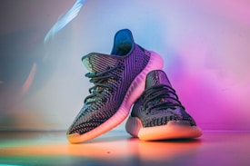 A pair of stylish, dark-colored sneakers with textured patterns, placed on a surface that reflects ambient rainbow-colored lighting. The shoes have a distinct sole design, prominent laces, and exhibit a modern, fashionable look.