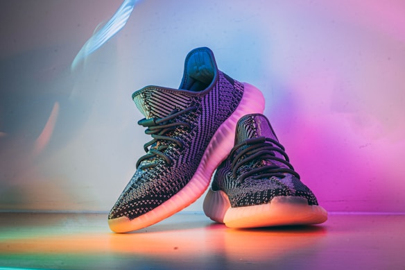 A pair of stylish, dark-colored sneakers with textured patterns, placed on a surface that reflects ambient rainbow-colored lighting. The shoes have a distinct sole design, prominent laces, and exhibit a modern, fashionable look.