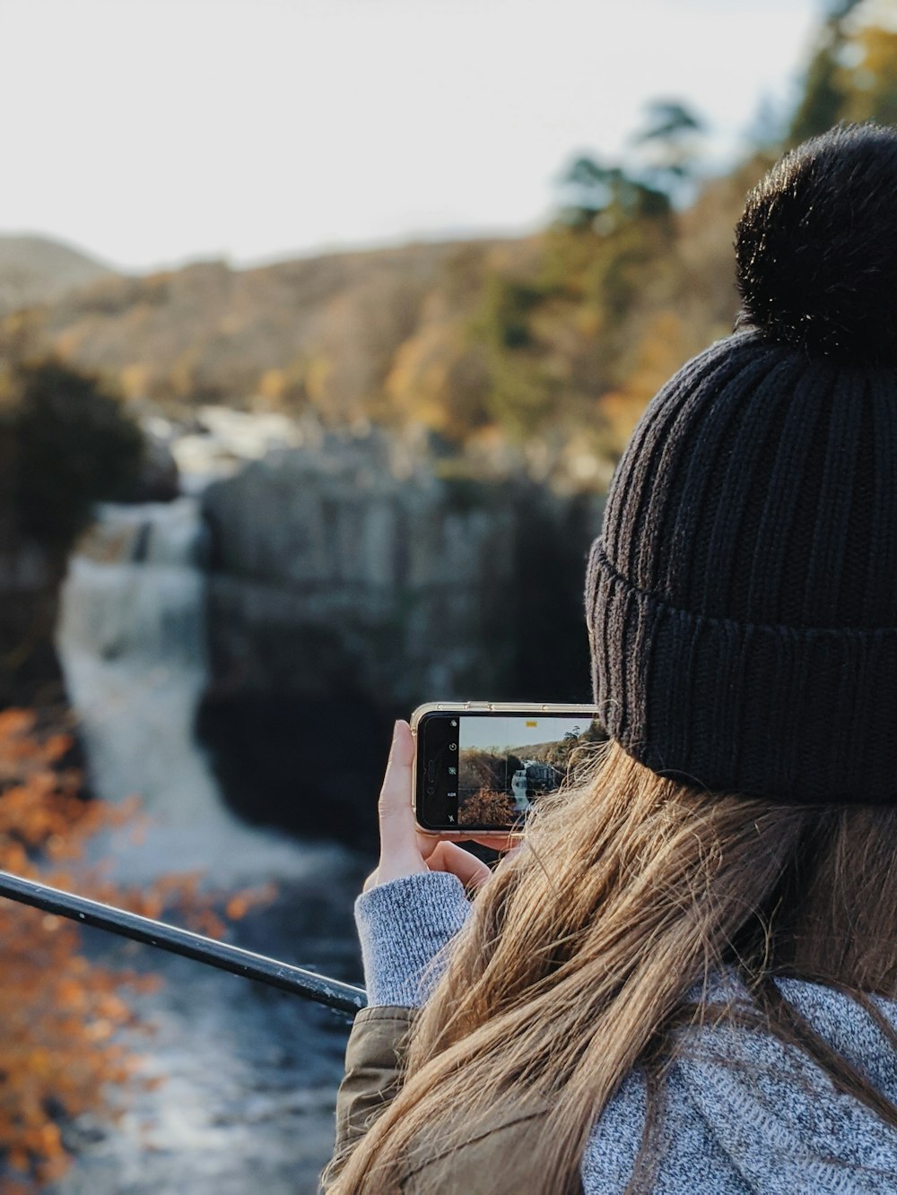 woman in black knit cap taking photo of waterfalls during daytime