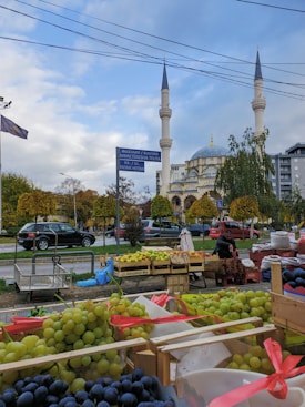 A street market with an assortment of fresh fruits, including green and black grapes, displayed in wooden crates. In the background, a mosque with intricate architecture and two tall minarets stands amidst trees with autumn foliage. Cars are parked along the street, and a vendor is arranging produce at a stall.