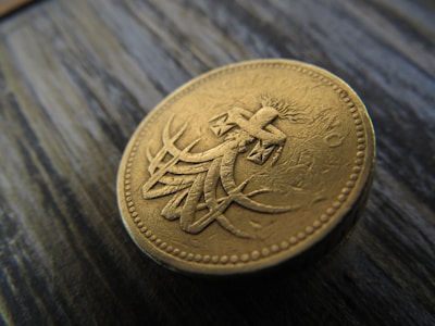 A close-up view of a gold-colored coin with intricate carvings and an embossed design featuring a stylized cross and abstract elements. The coin rests on a textured, dark, wooden surface.