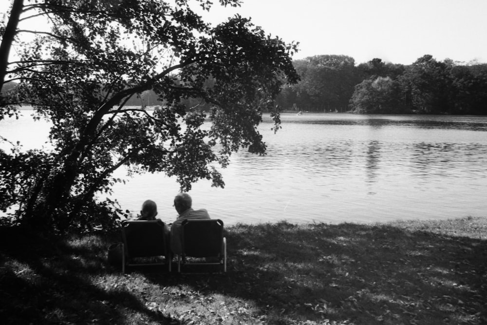 grayscale photo of 2 person sitting on chair near body of water