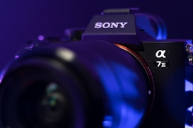 A close-up of a Sony Alpha 7 III camera, highlighting the camera body and lens in a moody, low-light setting. The focus is on the brand logo and model name, with a bluish-purple lighting effect giving the equipment a sleek and professional look.
