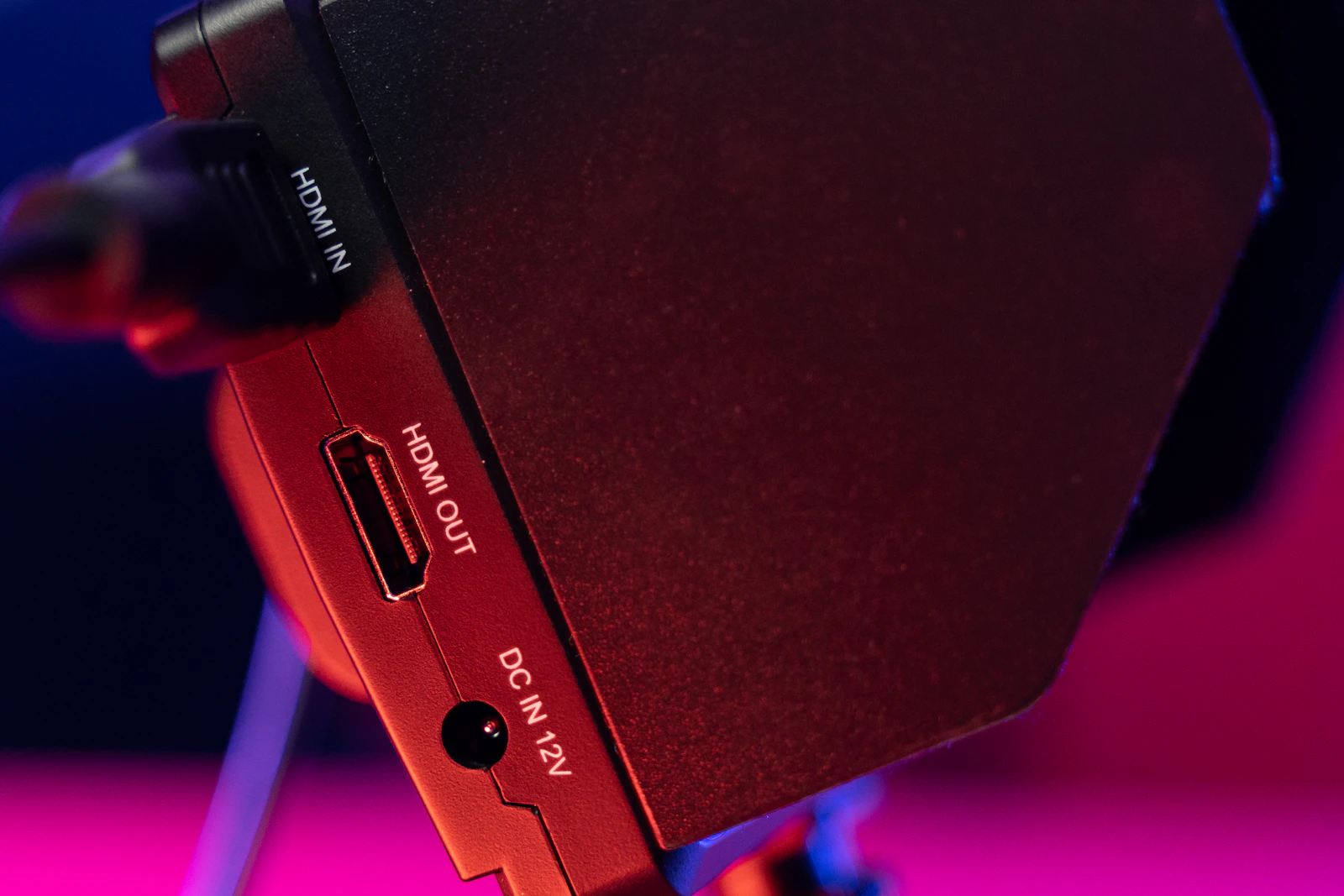 Close-up of a black device side panel with an HDMI cable plugged into HDMI IN, an empty HDMI OUT port below it, and a DC IN 12V jack, lit with red and pink light