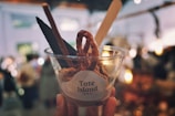 A clear plastic cup is filled with a dessert that includes a pretzel, a dark wafer, and a wooden stick. The cup has a label with 'Tate Island' written on it. In the background, there is a bokeh effect created by blurred lights and people, giving a warm and cozy ambiance.