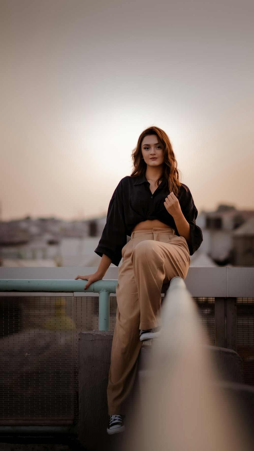 woman in black shirt and beige pants sitting on gray metal fence during daytime