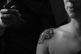 An intricate henna design is being applied to a person's shoulder. The scene is captured in black and white, highlighting the contrast between the skin and the detailed artwork. A hand is holding a cone, presumably filled with henna, delicately working on the design.