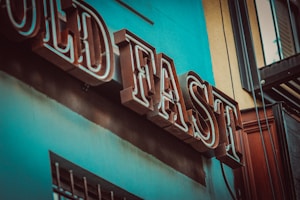 A vintage-style, bold block-lettered sign on a building that reads 'OLD FAST', with rich turquoise and earthy brown tones visible on the wall and sign itself.