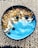 A bowl of vibrant blue smoothie garnished with granola, fresh blueberries, chia seeds, shavings of coconut, and other small seeds, placed on a woven mat.