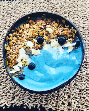 A bowl of vibrant blue smoothie garnished with granola, fresh blueberries, chia seeds, shavings of coconut, and other small seeds, placed on a woven mat.