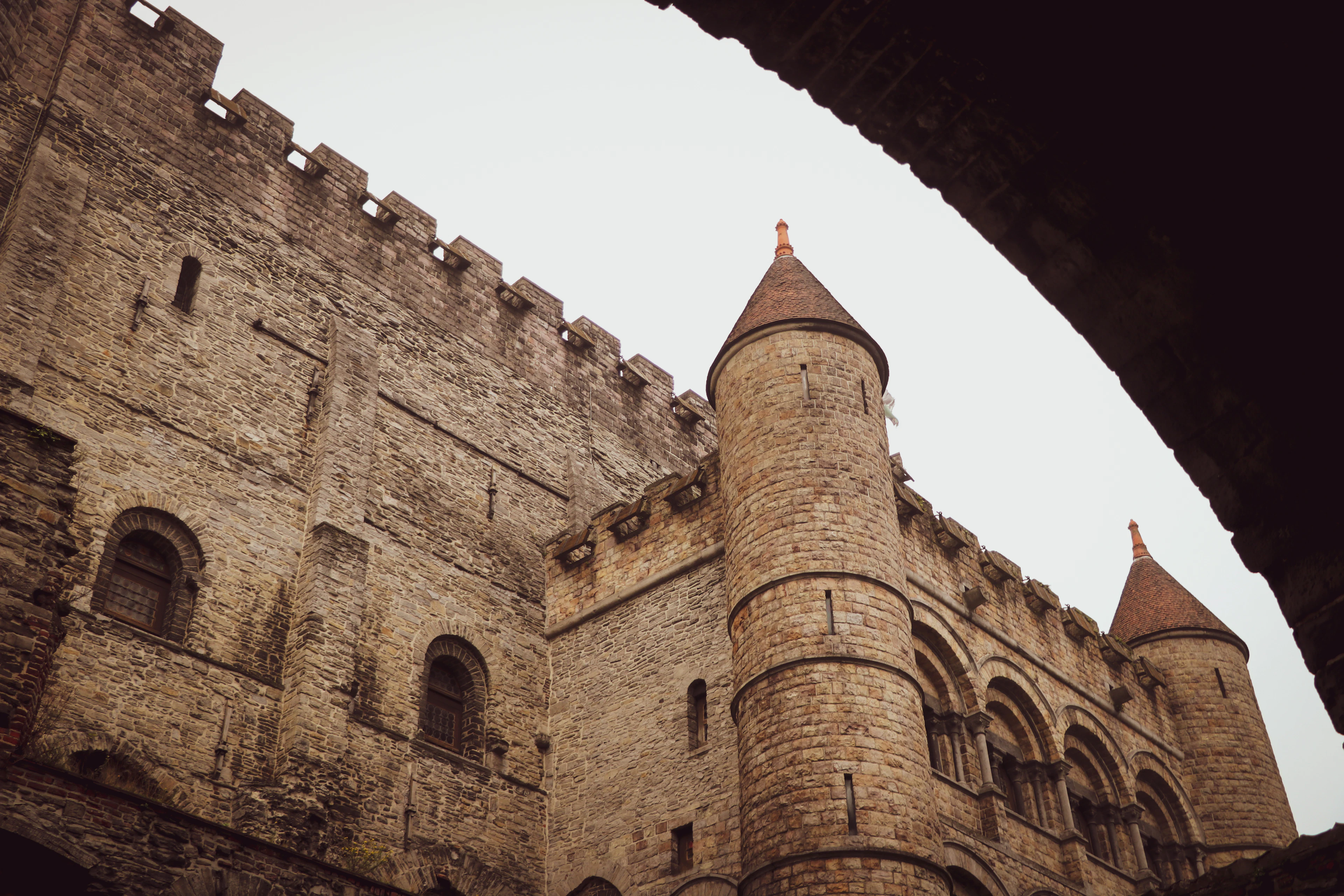 Looking up through a stone archway at the weathered turrets and battlements of Ghent's Gravensteen castle.
