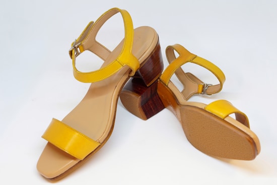 A pair of elegant yellow heeled sandals with leather straps and wooden block heels. The design is minimalistic, featuring a single buckle closure near the ankle. The sandals are positioned against a clean white background, highlighting their vibrant color and stylish design.