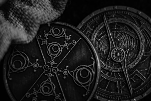 Two ornate, circular metallic objects with intricate geometric and celestial designs lie side by side, partially draped with a textured fabric. The patterns suggest celestial navigation or alchemical symbols.