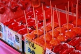 A colorful display of candied fruits, primarily featuring bright red treats on sticks arranged neatly in rows. Each piece is placed in a shiny, reflective red foil wrapper. The image includes signs with Japanese writing, suggesting a festive or market setting.