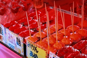 A colorful display of candied fruits, primarily featuring bright red treats on sticks arranged neatly in rows. Each piece is placed in a shiny, reflective red foil wrapper. The image includes signs with Japanese writing, suggesting a festive or market setting.