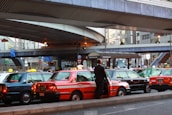 A line of taxis, each with unique color schemes, is parked under a large, elevated roadway in a busy urban setting. A man stands beside one of the taxis, which has a distinct orange stripe pattern. Surrounding infrastructure includes modern buildings with large windows and various signage.